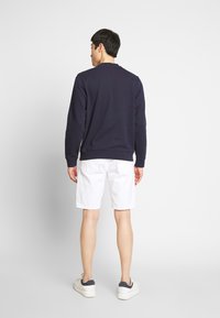 Felpa navale con maniche lunghe, scollatura rotonda e polsini a costine, abbinata a pantaloni corti bianchi e sneaker con accenti grigi. Sfondo semplice.