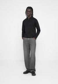 Μαύρο πόλο με μακριά μανίκια και φερμουάρ στον γιακά, συνδυασμένο με φαρδιά γκρι τζιν και μαύρα παπούτσια. Το ύφασμα φαίνεται απαλό και καθημερινό.