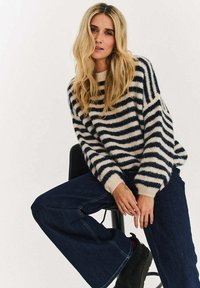 Pasiasty sweter w beżowo-granatowe paski, o puszystej fakturze, szerokich rękawach i ściągaczach, zestawiony z szerokimi spodniami z ciemnego denimu.