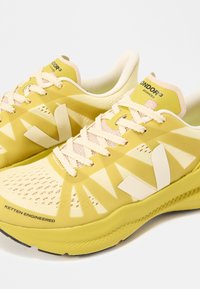 Κίτρινα και εκρού ελαφριά παπούτσια τρεξίματος με λευκά κορδόνια, με λογότυπα "V" και κείμενο που γράφει "Ketten Engineered" και "Condor 3 Advanced".