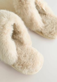 Pantofle z umělé kožešiny v světlé béžové barvě s plush, měkkou texturou. Design zahrnuje otevřenou špičku a měkkou vycpanou podrážku.