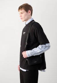 Crna crossbody torba s teksturiranom površinom, srebrnim hardverom i podesivim remenom. Nosiva preko crne majice i prugaste košulje.
