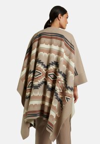 Beiger Poncho mit gemustertem Design in Braun, Creme und Marineblau, mit tribalischen Motiven und einem lässigen Schnitt, aus weichem Stoff gefertigt.