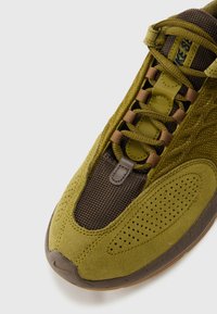 Sapatilhas verde-oliva com um upper em tecido texturizado, painéis em malha castanha, ilhós reforçados e detalhes perfurados nas laterais.