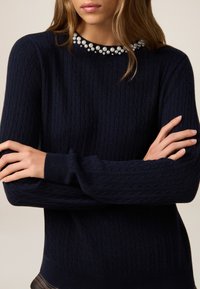 Maglione a trecce blu navy con scollatura rotonda decorata con perle. Maniche lunghe con polsini a coste. Texture liscia su tutto il capo.