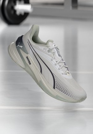 Chaussure de course Puma gris clair et blanc avec logo noir et détails, présentée flottant au-dessus d'un sol brillant dans une salle de sport.
