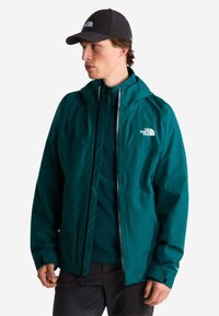 Άνδρας που φοράει πετρόλ μπουφάν The North Face με κουκούλα και μαύρο καπέλο κοιτάζει αριστερά σε απλό φόντο.