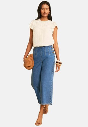 Frau trägt eine weiße, strukturierte Kurzarmbluse, hoch taillierte blaue Jeans mit weitem Bein und seitlichen Knöpfen, hält eine braune Tasche und trägt Sandalen.