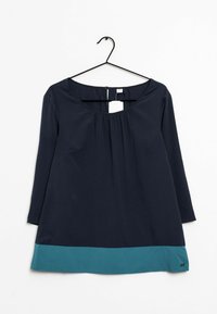 Top azul marino con mangas tres cuartos, escote con pliegues y un dobladillo de color verde agua en la parte inferior. Cuelga de un gancho negro.