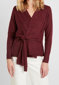 Femme portant un pull cache-cœur bordeaux à manches longues noué à la taille, associé à un pantalon blanc taille haute sur un fond uni.