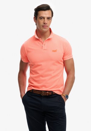 Mand iført laksefarvet poloshirt, mørke bukser, fletbælte i brun farve og armbåndsur, står med hænderne i lommerne.