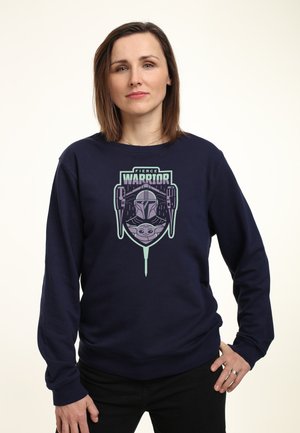 Kobieta z krótkimi brązowymi włosami, ubrana w granatową bluzę z napisem "Fierce Warrior" oraz grafikami Mandalorianina i Baby Yody, stoi z jedną ręką na biodrze.