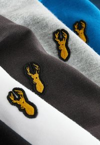 Drei Stoffstücke in Blau, Grau und Dunkelbraun, jeweils mit einem gelben gestickten Hirschlogo auf schwarzem Hintergrund. Weiche Textur.