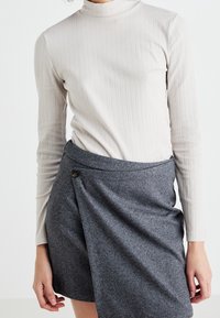 Langärmliges, geripptes beiges Oberteil, kombiniert mit einem grauen Herringbone-Wickelrock, der eine Knopfdetail an der Taille hat.