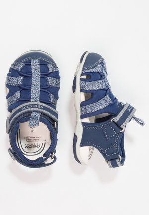 Navy blue sandals with a mesh upper, elastic straps, and a Velcro closure. Features a white rubber sole and contrasting stitching accents.