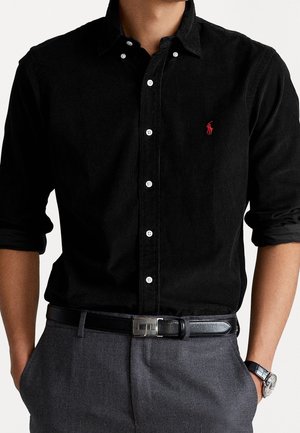 Homme portant une chemise noire à boutons avec un logo brodé rouge, un pantalon gris, une ceinture noire et une montre-bracelet, les mains dans les poches.