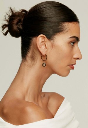 Pendiente de aro dorado con un charm circular de intrincados detalles. Piel suave, cabello recogido en un moño bajo, vistiendo una blusa de hombros descubiertos.