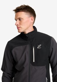 Ung man med kort hår iförd en svart och mörkgrå zip-up jacka med en vit dragkedjeficka och logotyp på bröstet, som tittar åt sidan.