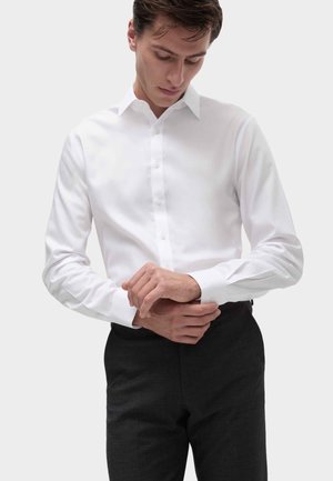 Witte lange mouwen shirt met een knopenfront, klassieke kraag en manchetten, gecombineerd met donkere op maat gemaakte broek, vastgelegd tegen een neutrale achtergrond.