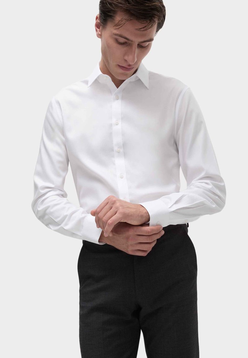 Witte lange mouwen shirt met een knopenfront, klassieke kraag en manchetten, gecombineerd met donkere op maat gemaakte broek, vastgelegd tegen een neutrale achtergrond.