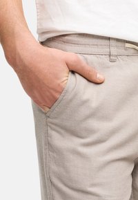 Hanit beige-housut, joissa on tekstuuri, sivutaskut ja siisti vyötärö. Käsi lepäilee taskussa, esitellen kankaan yksityiskohtia.