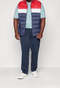 Rote und marineblaue Steppweste über einem hellblauen Hemd, kombiniert mit marineblauen Hosen und weißen Sneakers. Steppdesign mit horizontaler Naht.