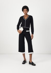 Maglione nero a scollo a V con piping bianco, abbinato a culotte nere a trama con risvolti bianchi e mocassini neri. Borsa a tracolla in mano.