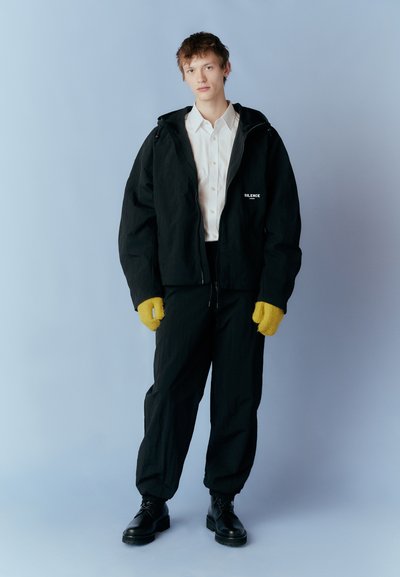 Black hooded jacket with a zip, white shirt, black trousers, yellow gloves, and black boots, standing against a pale blue background.