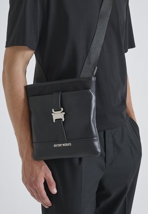Borsa a tracolla nera in pelle con una forma strutturata, chiusura in metallo argentato e nome del brand stampato sul fondo. Comprende una tracolla regolabile.