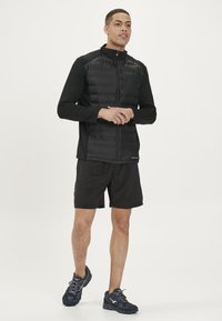 Schwarze gepolsterte Weste mit hohem Kragen und Reißverschluss vorne, getragen über einem schwarzen Langarmshirt. Kombiniert mit schwarzen Shorts und dunklen Sportschuhen.