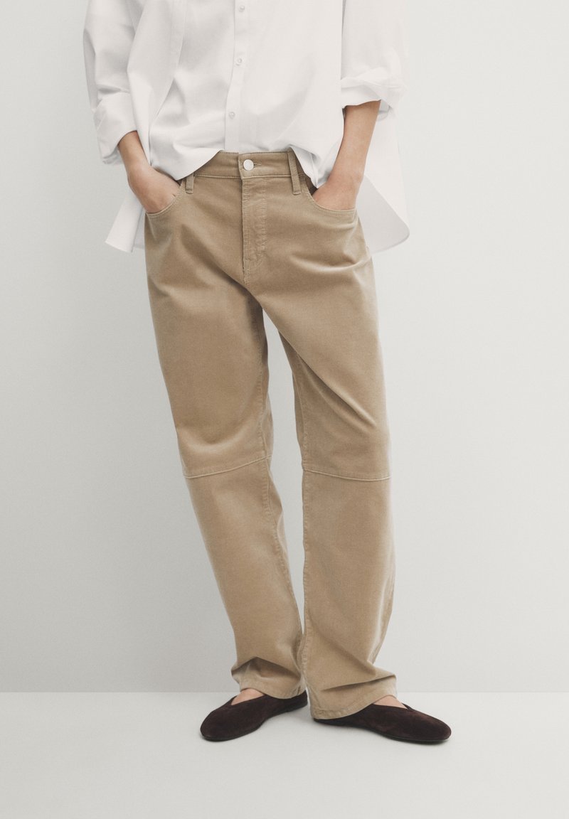 Personne portant un pantalon beige à jambe droite, une chemise blanche boutonnée avec les manches retroussées, les mains dans les poches, et des chaussures plates marron foncé.