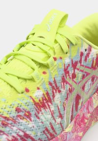 Heller gelber Sportschuh mit einem strukturierten, gemusterten Obermaterial, das pinke und blaue Akzente aufweist, sowie einem Logo-Tag auf der Zunge.
