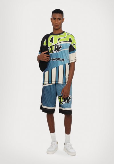 Maglietta a maniche corte multicolore e pantaloni corti abbinati in blu, con motivi geometrici, loghi e un design sportivo. Sneakers bianche indossate.