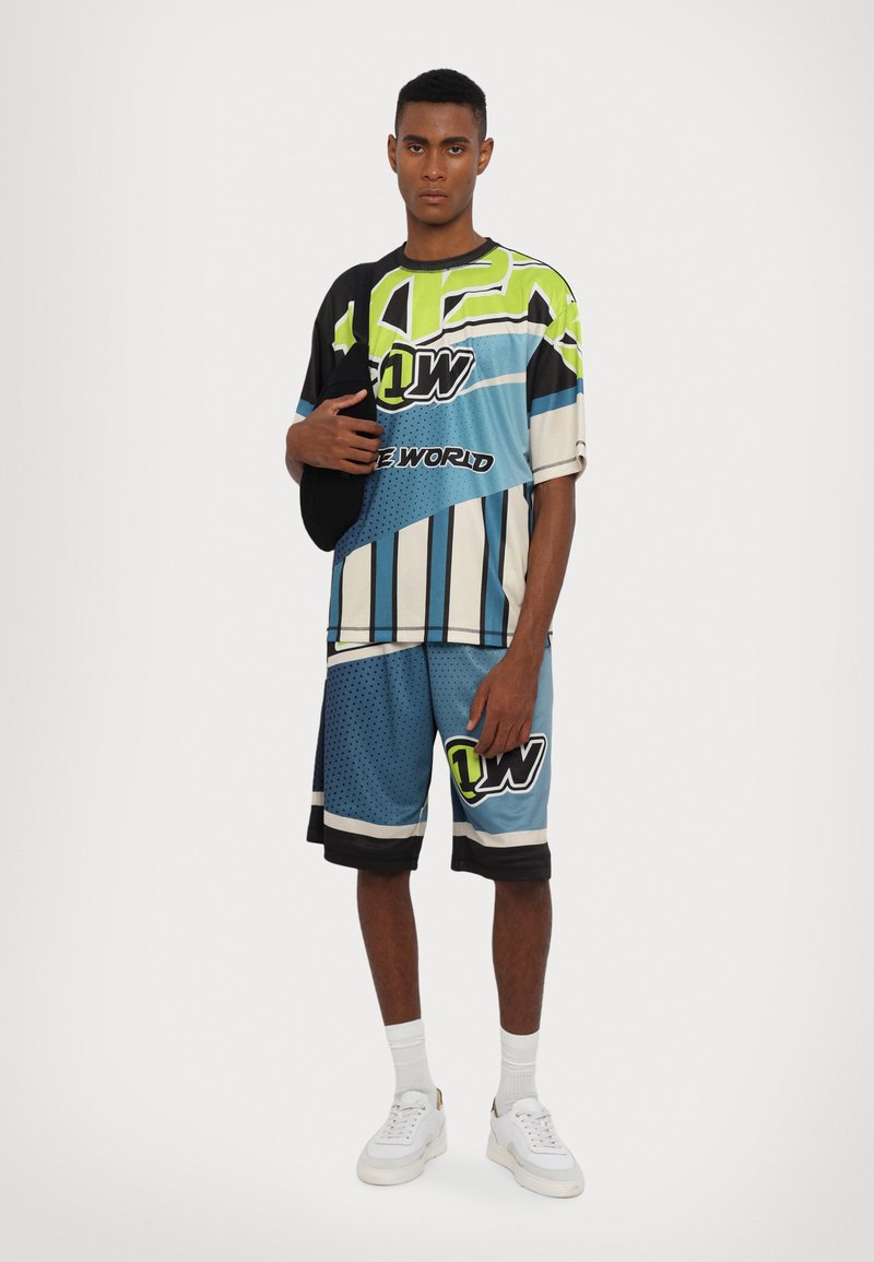 Tricou cu mânecă scurtă, în nuanțe multiple, și pantaloni scurți asortati de culoare albastră, cu modele geometrice, logo-uri și un design sportiv. Se poartă adida�și albi.