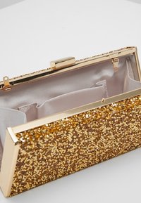 Błyszcząca kopertówka w kształcie trójkąta, z złotym metalowym zapięciem i gładkim, satynowym wnętrzem z podzielonymi przegródkami.