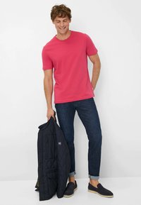 Outfit da uomo composto da una t-shirt rosa brillante, jeans di denim blu scuro e una giacca trapuntata nera. Gli accessori includono scarpe slip-on scure.