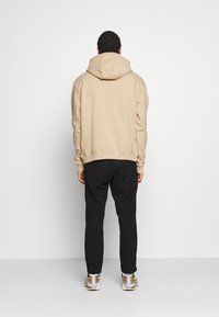 Felpa beige con cappuccio e vestibilità comoda, con polsini a costine, abbinata a pantaloni neri e scarpe sportive con dettagli dorati.