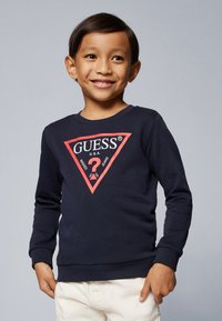 Mørkeblå hettegenser med lange ermer, med et rødt trekantet logo med teksten "GUESS U.S.A." og et spørsmålstegn i midten.