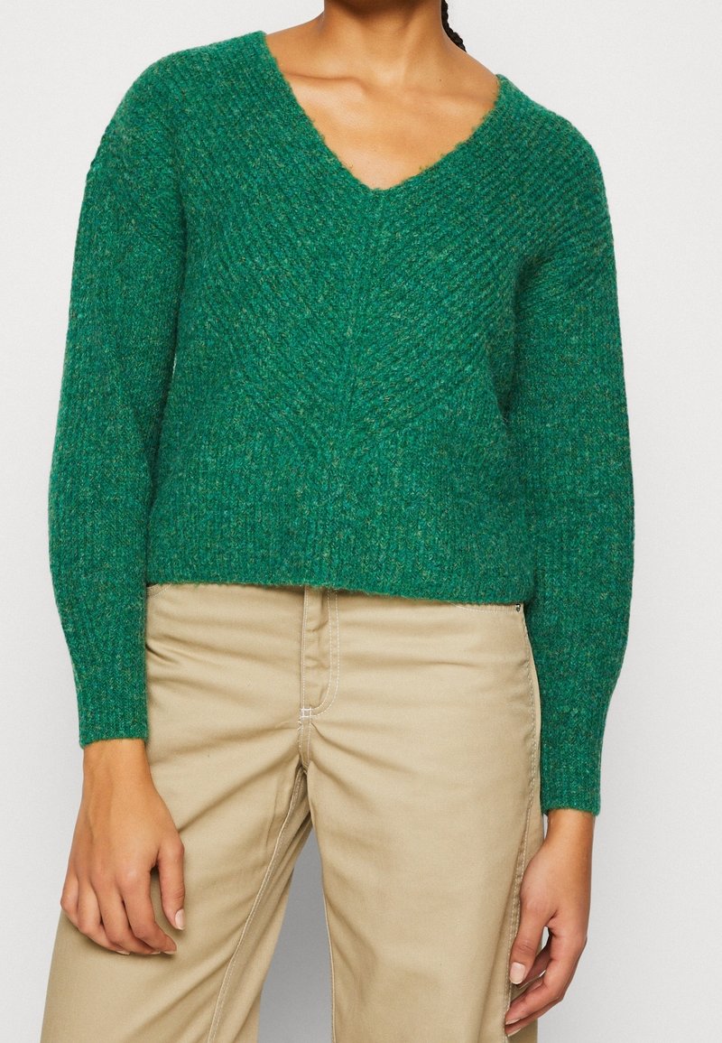 Personne portant un pull vert texturé à col en V et un pantalon beige, debout les bras détendus le long du corps sur un fond uni.