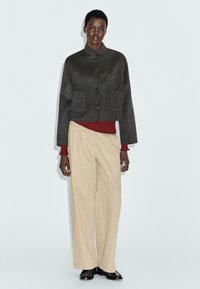Modella che indossa un giubbotto grigio scuro con bottoni, una maglietta a righe rossa e nera, pantaloni beige a gamba larga e scarpe nere su uno sfondo chiaro e semplice.