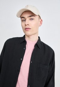 Camicia nera a maniche lunghe sopra una maglietta rosa chiaro, con un berretto beige recante un logo, su uno sfondo neutro.