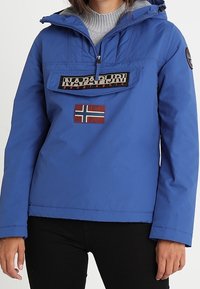 Modrá bunda s kapucí, předním zipem a klopnou kapsou s nášivkou vlajky Norska a štítkem značky "Napapijri".