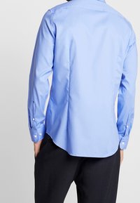 Ljusblå långärmad skjorta med knappmanschetter, slät textur, rundad nederkant och klassisk krage. Bakvy visar skräddarsydd passform och sömdetaljer.