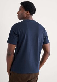 Tamsiai mėlynos spalvos medvilninis marškinėliai su apvaliu kaklu ir trumpomis rankovėmis, turintys lygią tekstūrą ir laisvą siluetą, matomi iš nugaros.