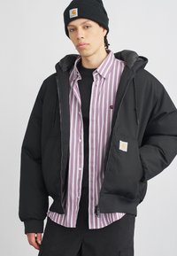 Mlad moški v črni kapa, črni pufast jakni, vijolični in beli prugasti srajci s gumbi ter črnih hlačah na enobarvni podlagi.