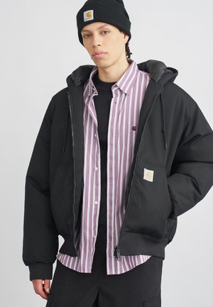 Joven vistiendo un gorro negro, una chaqueta de plumas negra, una camisa de botones a rayas moradas y blancas, y pantalones negros contra un fondo sencillo.