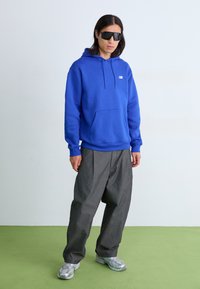 Blå hettegenser med kengurulomme, kombinert med løse grå bukser og sølvfargede sneakers. Modellen har på seg svarte solbriller.