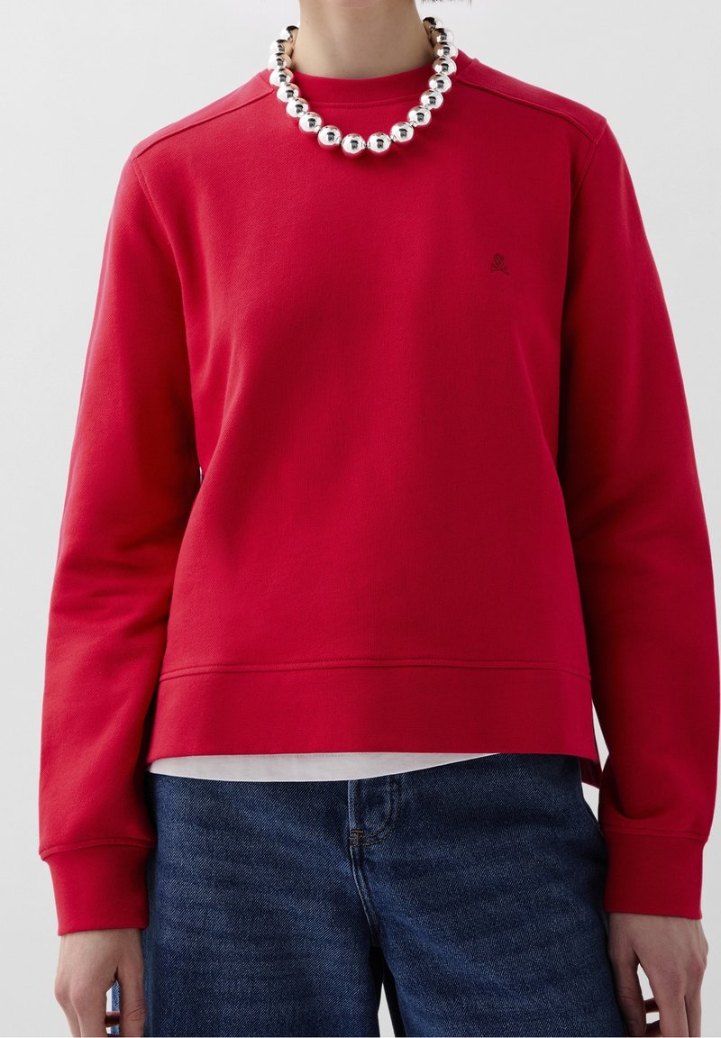 Personne portant un sweat-shirt rouge à manches longues, un jean bleu, un sous-vêtement blanc et un collier épais en perles argentées, sur un fond gris uni.
