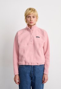 Persoon in een roze Columbia fleecetruien met drukknoopjes en blauwe spijkerbroeken, staande tegen een effen witte achtergrond.