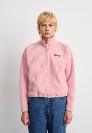 Person wearing a pink Columbia fleece pullover with snap buttons and blue jeans, standing against a plain white background.